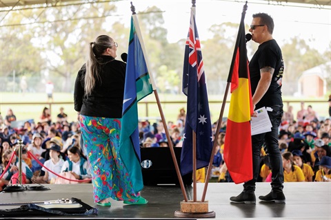 Two people on stage with First Nations Flags addressing school students